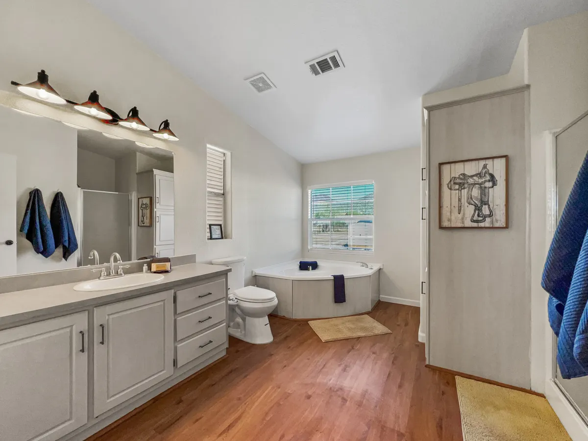 A spacious bathroom featuring a deep soaker bathtub, vanity with a mirror, window letting in natural light, decorative artwork, and neatly folded towels. The ambiance is serene and spa-like, perfect for unwinding.