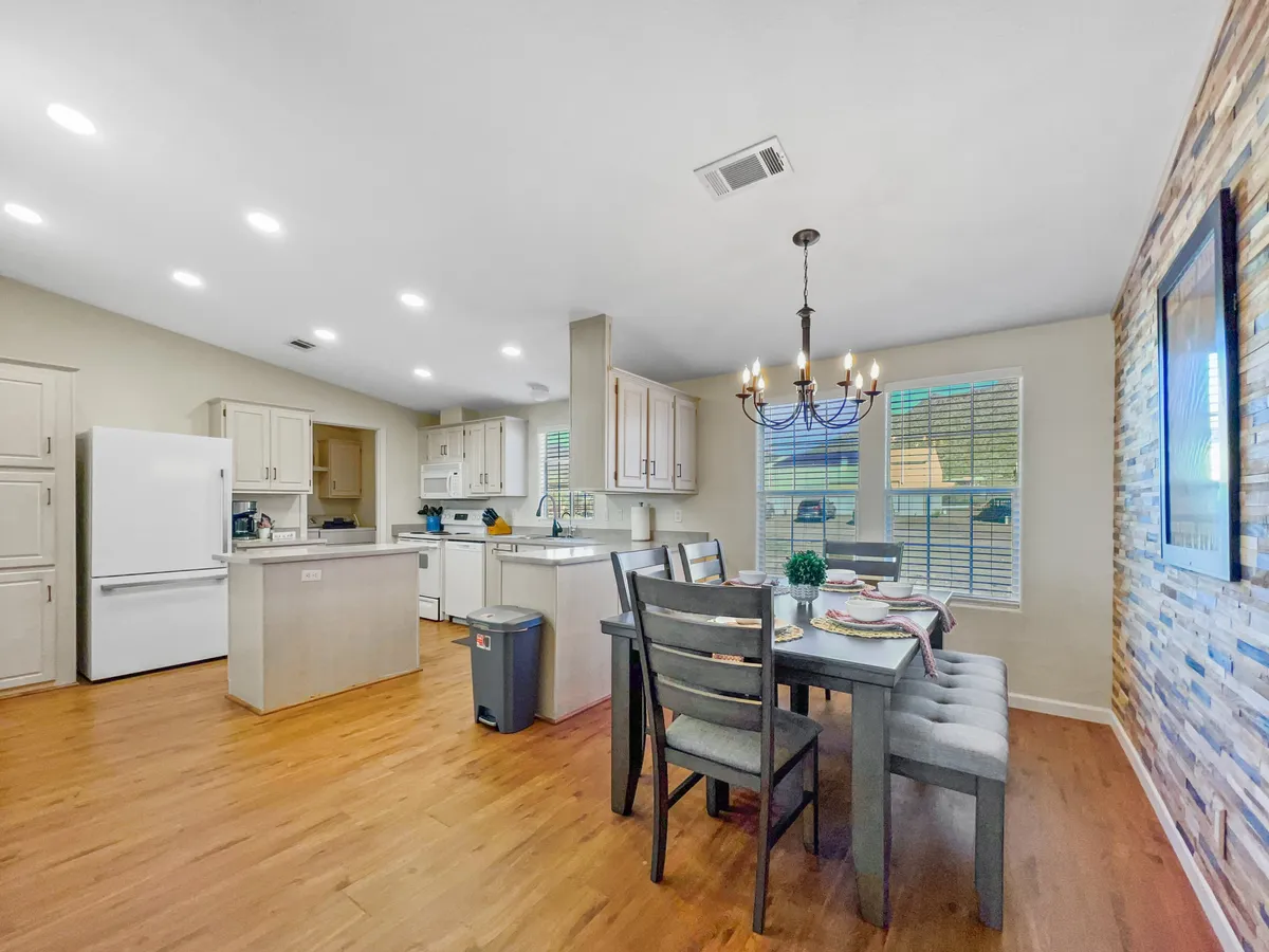 This image showcases a warm, inviting kitchen and dining space featuring a kitchen island, an elegant chandelier, a sturdy dining table, and an exposed stone wall. The scene is bathed in natural light, emphasizing the room’s welcoming ambiance and the multiple areas available for prepping, cooking, and gathering.