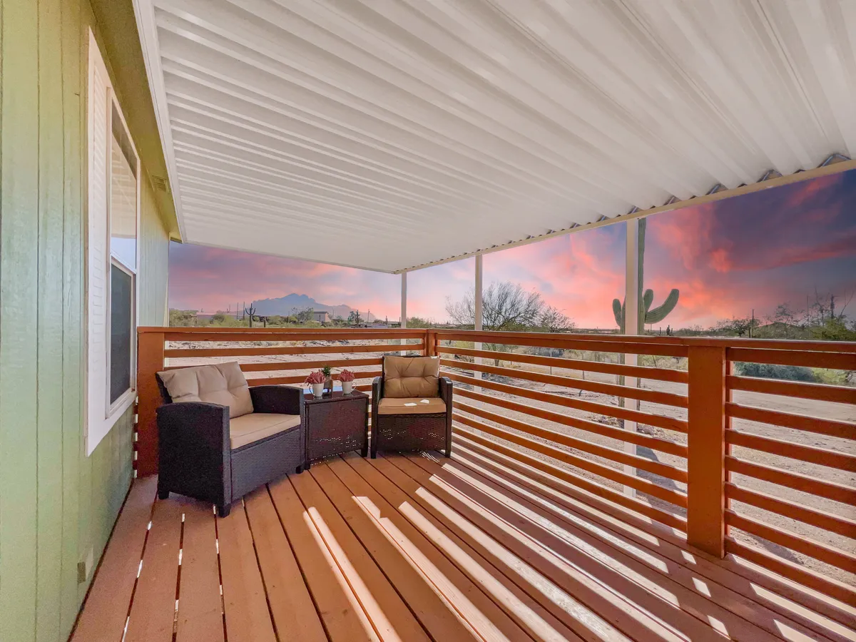 An inviting outdoor seating area with a wooden railing overlooks a stunning mountain view and expansive desert landscape, all glowing under a vibrant sunset. The setup suggests a seamless transition from focused work to moments of reflection or creative inspiration, with nature just outside the window.