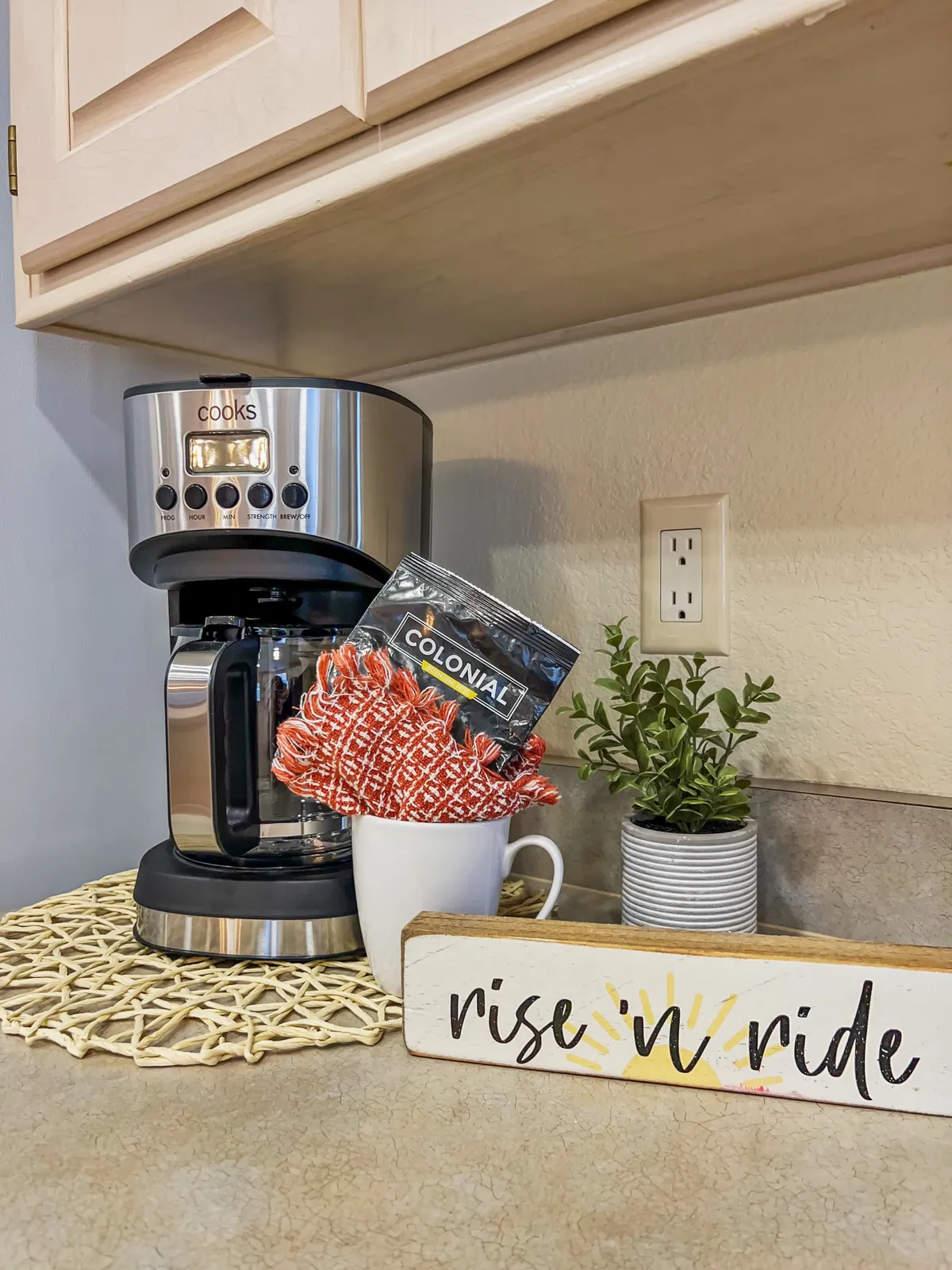 A cozy kitchen corner featuring a coffee maker, a mug with a dish towel, a decorative sign, and a potted plant. The space is bathed in natural light and exudes a welcoming atmosphere, ideal for morning routines or coffee breaks.