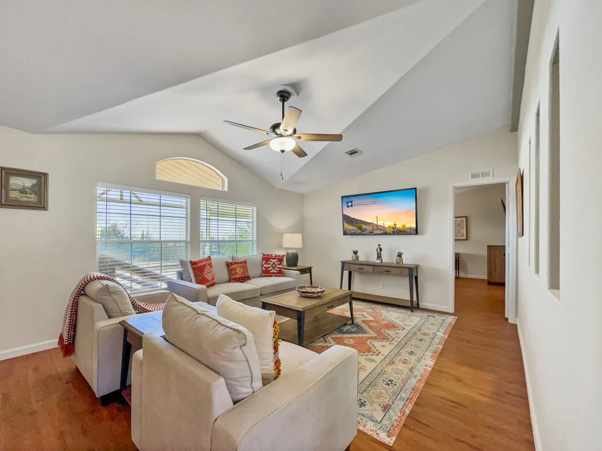 This image showcases a cozy living room featuring a sofa set, coffee table, and a television. Large windows with blinds allow natural light to filter in, and the arrangement encourages relaxing with family, entertaining guests, or hosting movie nights. The warm, comfortable mood is accentuated by inviting decor.