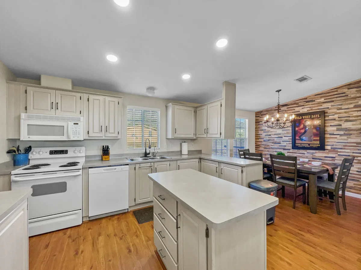 The image centers on a beautifully arranged kitchen and dining area featuring a spacious kitchen island with modern appliances, a wooden dining table with chairs, and a decorative wall. The overall mood is cozy and inviting, suggesting the perfect spot for both everyday meals and special gatherings.