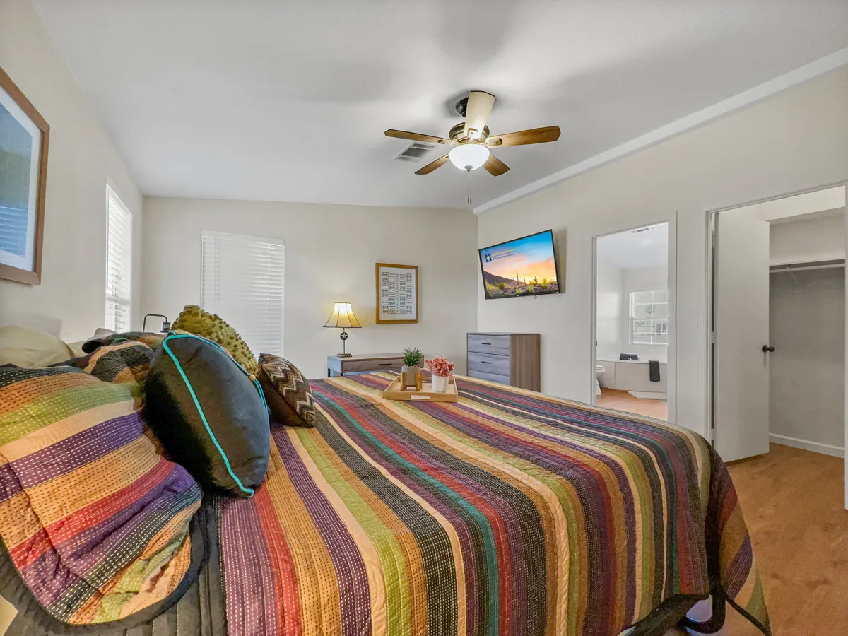 A bright and inviting bedroom featuring a colorful bedspread, ceiling fan, wall-mounted TV, nightstand with a lamp, and framed artwork. The room exudes comfort and warmth, ideal for unwinding after a day of adventure.